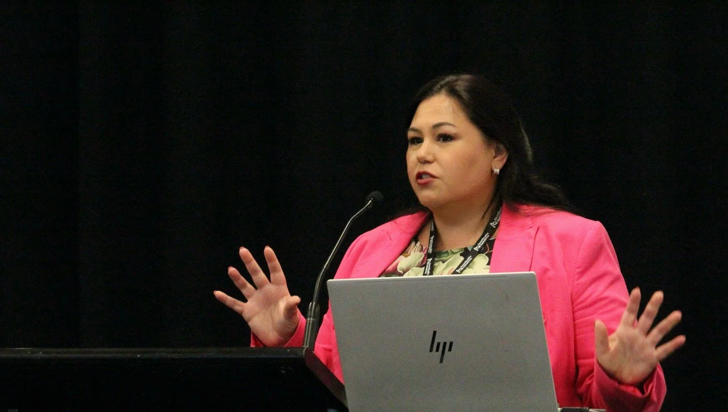 A woman with dark hair wearing a pink blazer and patterned blouse speaking at a podium with a microphone and a gray HP laptop in front of her against a black background.