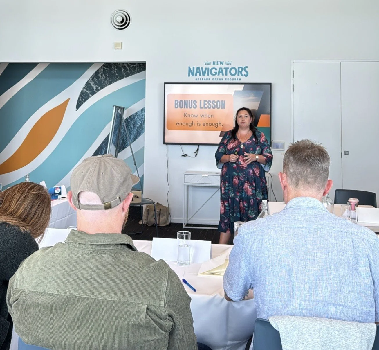 A woman is giving a presentation to a group of people in a conference room. She is standing in front of a screen that displays 'BONUS LESSON' and 'Know when enough is enough.' The room is decorated with a geometric mural on the wall, and the audience members are sitting around tables with glasses of water.