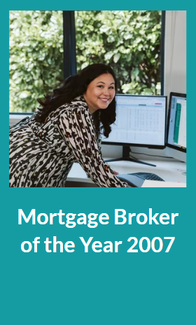 A woman in a patterned blouse working at a desk with dual computer monitors, smiling and looking at the camera, in front of a large window with greenery outside. Text reads 'Mortgage Broker of the Year 2007'.