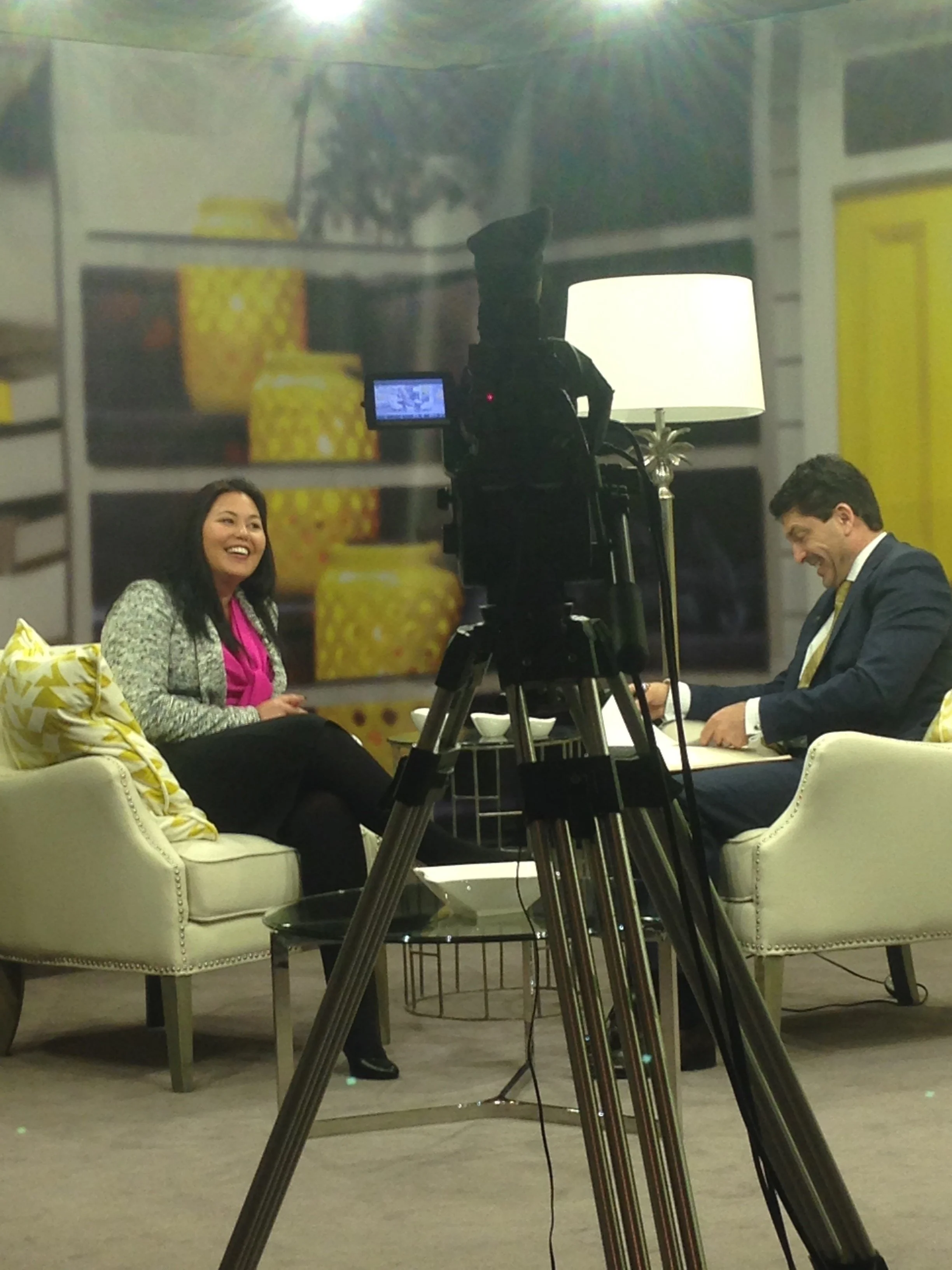Two people, a woman and a man, are sitting on cream-colored sofas in a room with a large camera on a tripod recording or broadcasting their conversation. The woman is smiling, the man is laughing. There is a lamp and a decorative background with yellow vases.