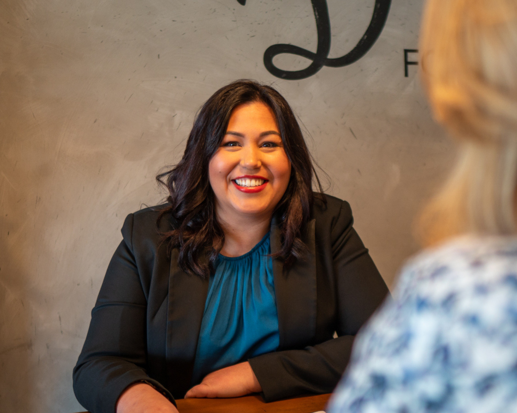 A woman with dark wavy hair, wearing a black blazer and blue blouse, smiling while talking to another woman with blonde hair in a patterned top, in an indoor setting.