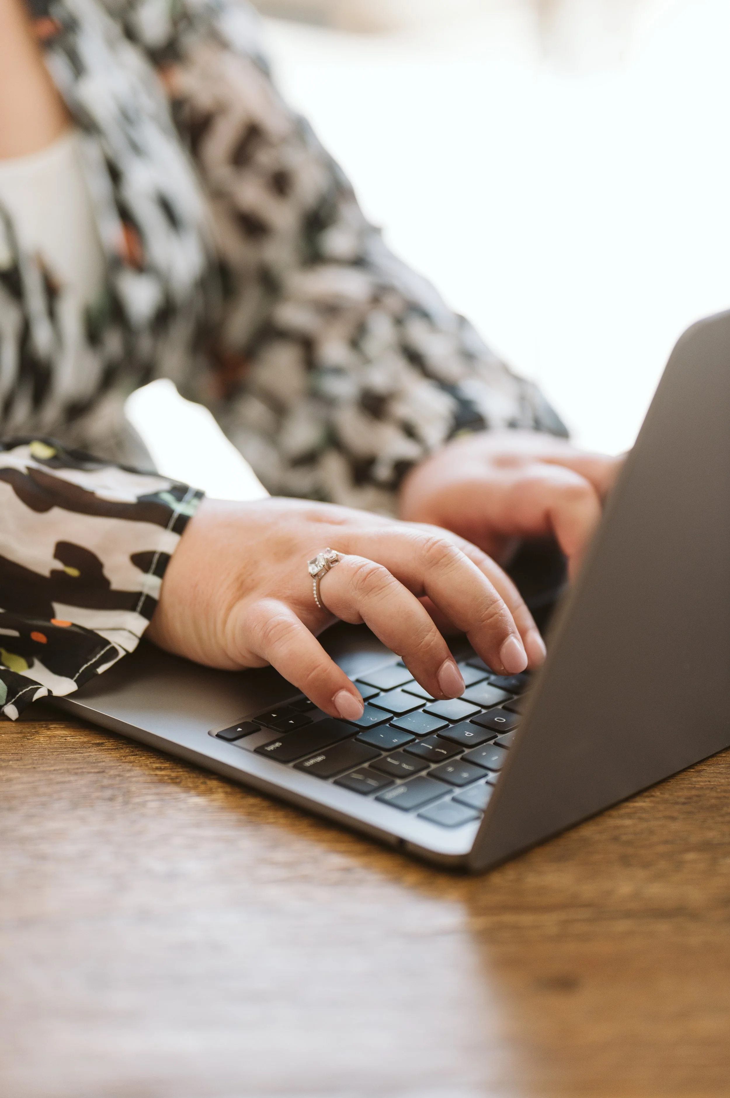 Close-up of a Amy Po-Ching typing on a laptop keyboard, wearing a patterned long-sleeve shirt, with a diamond ring on their finger, sitting at a wooden table.