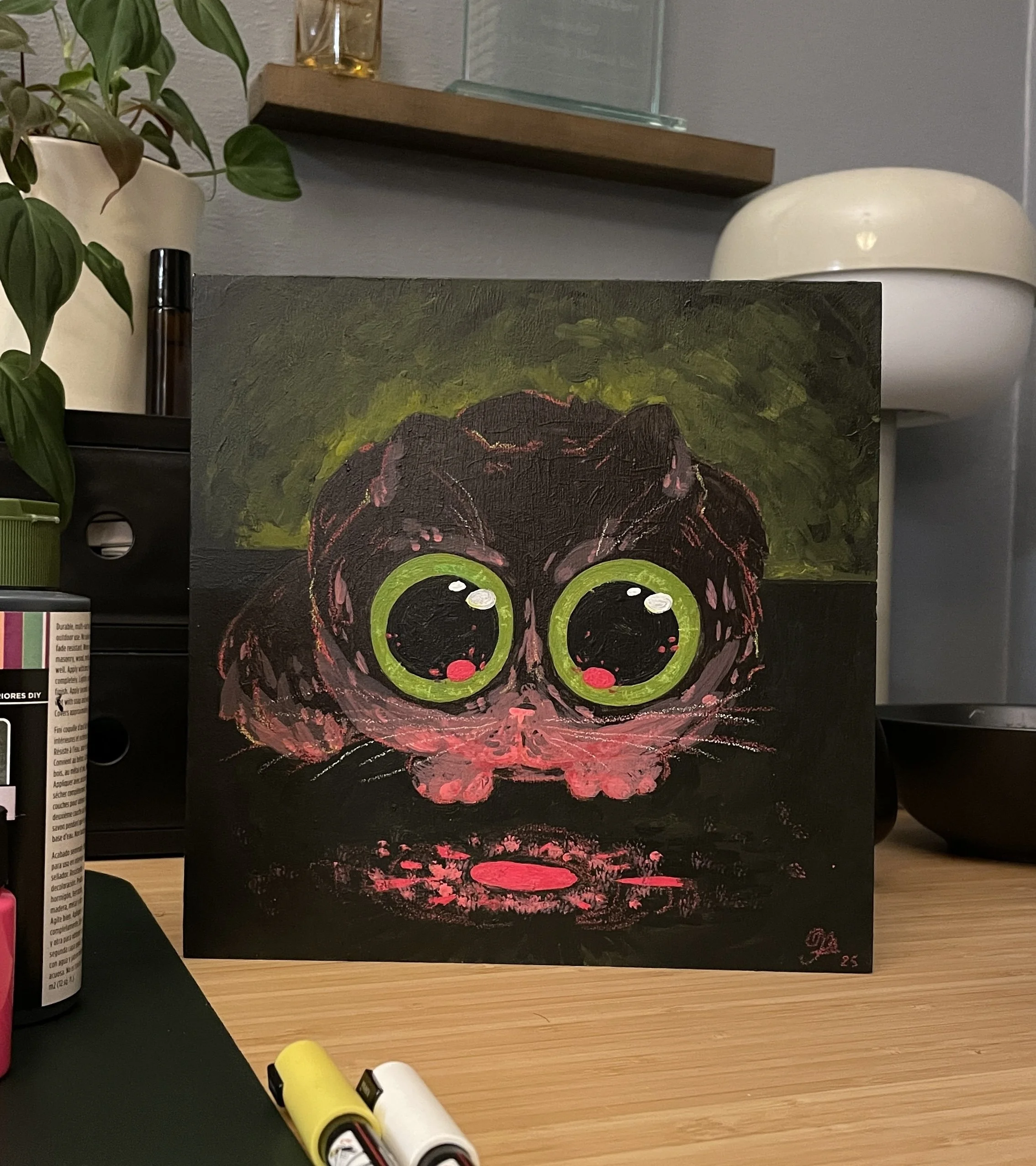 A painting of a cartoonish black cat with large green eyes and pink accents, resting on a black background. The painting is on a small canvas on a wooden table, with a plant, a white lamp, and a shelf with glass items in the background.