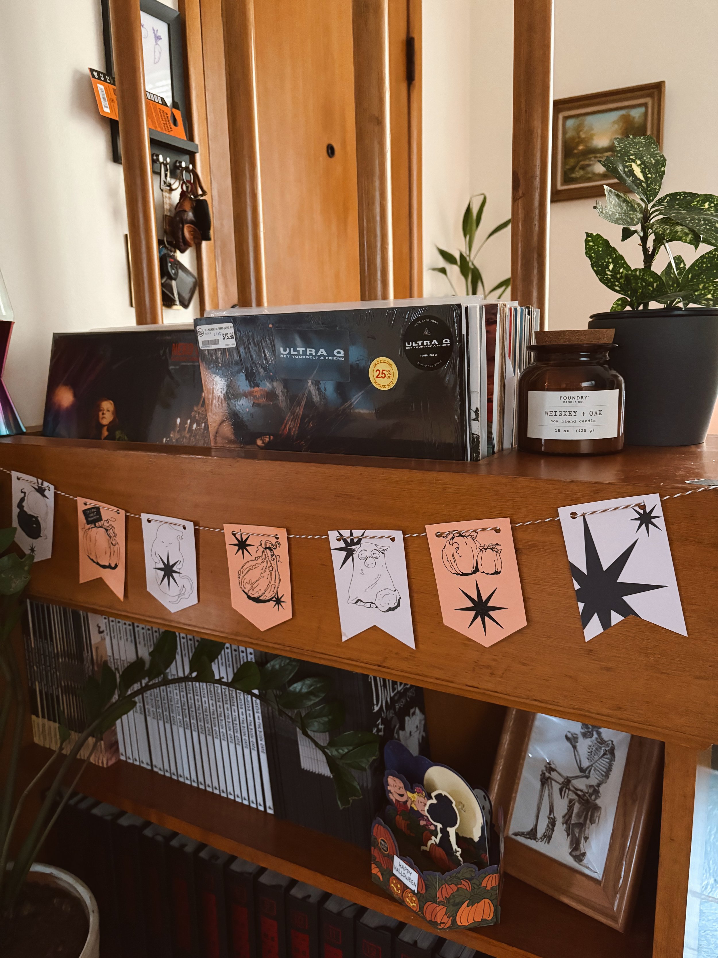 A wooden bookshelf with various items, including Halloween-themed decorations like a pumpkin garland, a 'Happy Halloween' sign, and a mouse figure, with a small table and potted plant nearby.