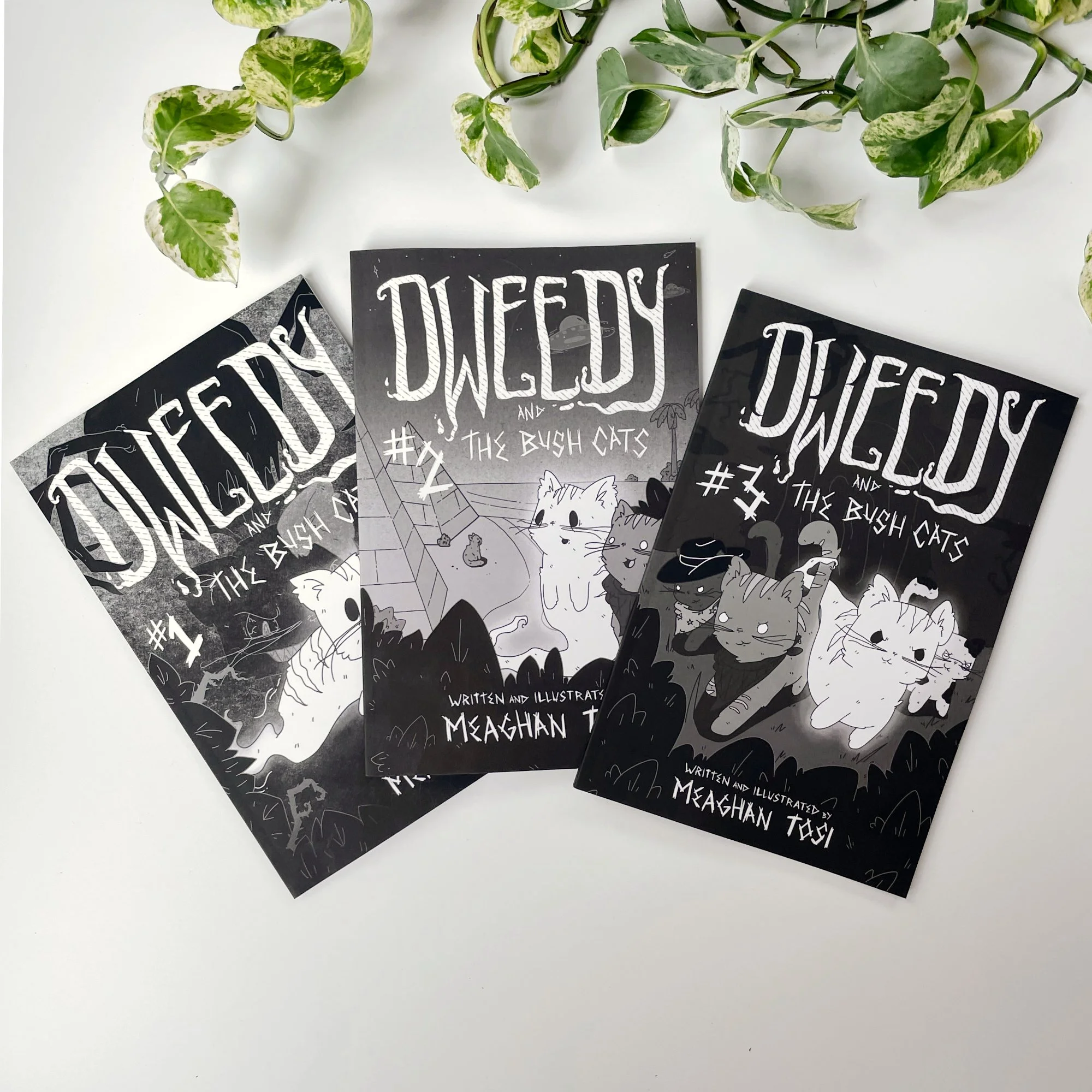 Black and white illustrated flyers with the text '#2 THE BUSH CATS' and 'WRITTEN AND ILLUSTRATED BY MEAGHAN T', surrounded by drawings of cats, houses, trees, and nature beneath a sprig of green and variegated leaves on a white background.