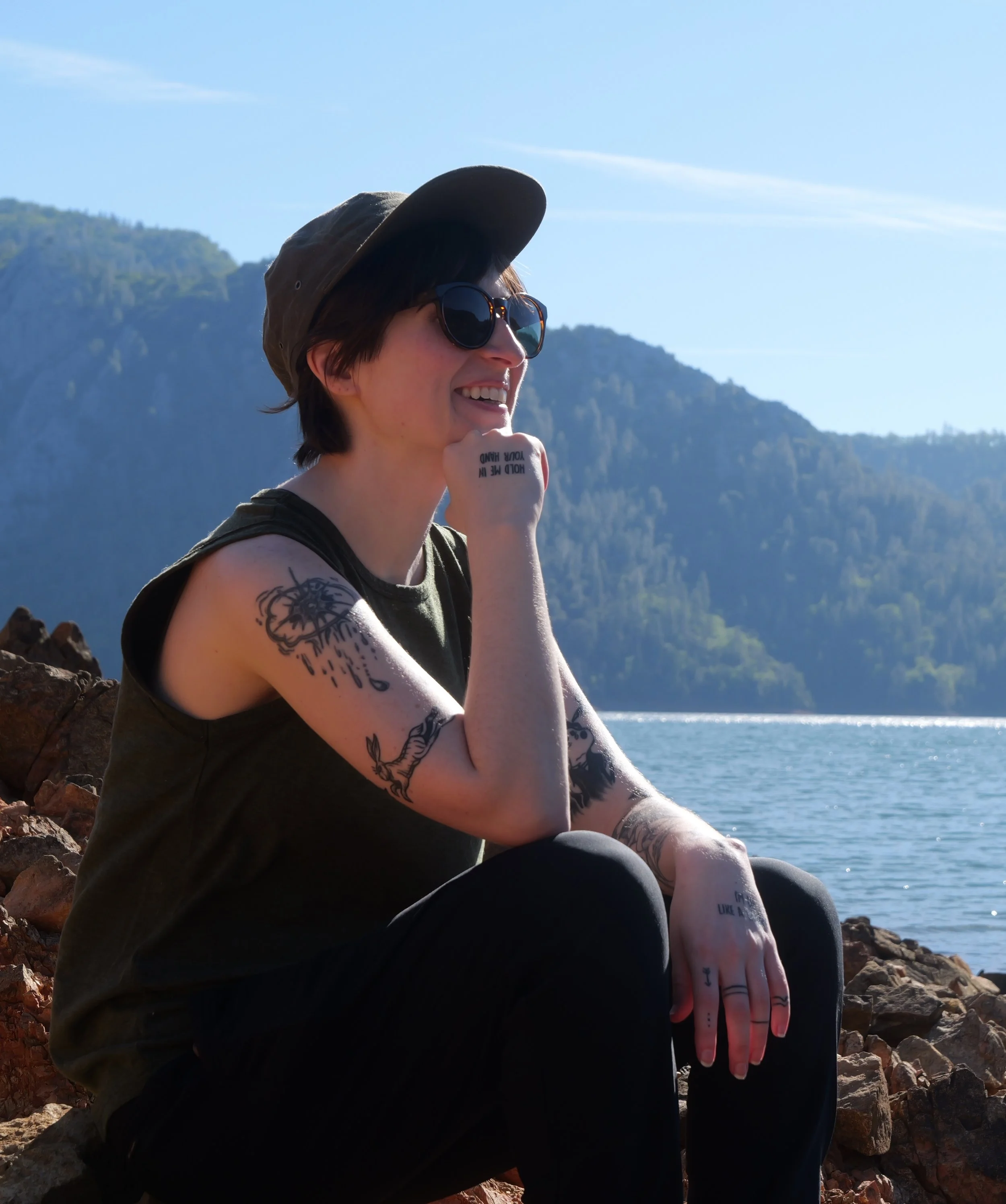 Person wearing sunglasses and a wide-brim hat, sitting on rocks by a body of water with mountain scenery in the background, smiling and resting chin on fist.