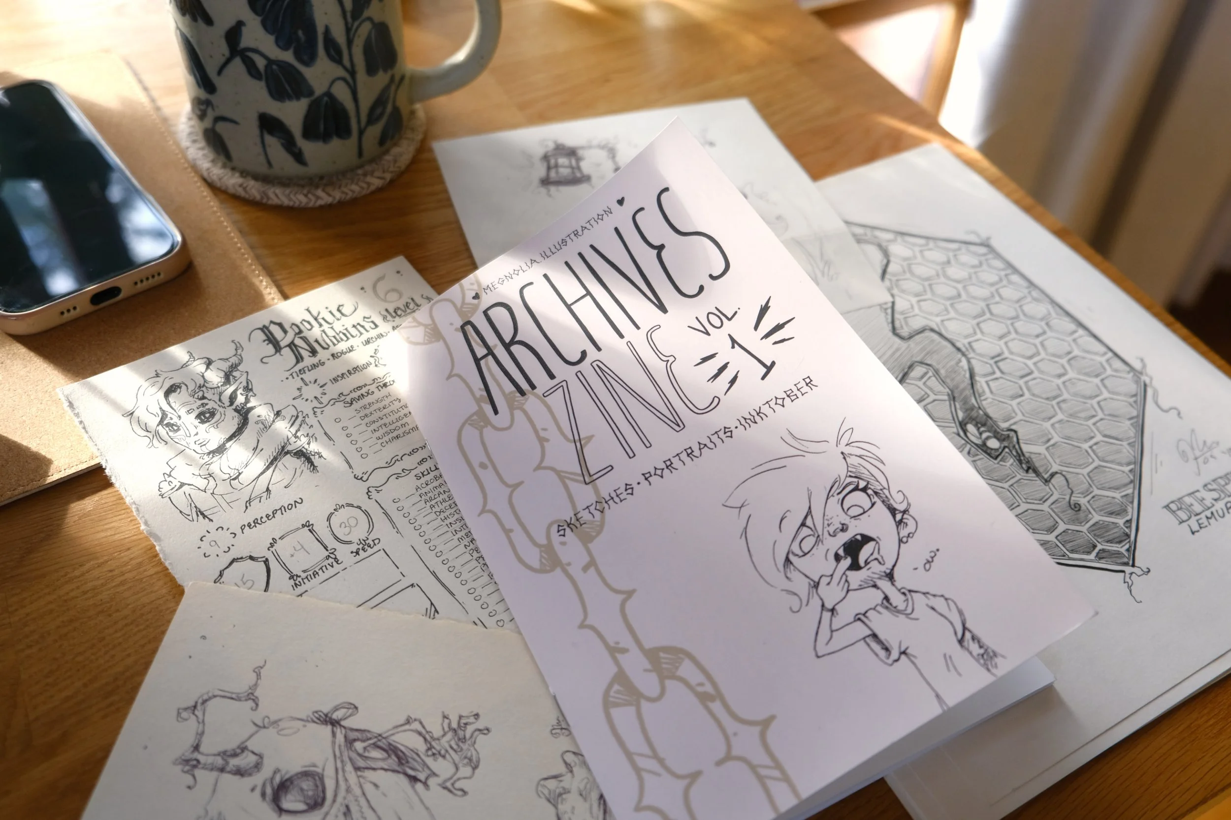 A wooden table with various comic sketches and illustrations, a smartphone, a mug with a floral pattern, and some printed papers.