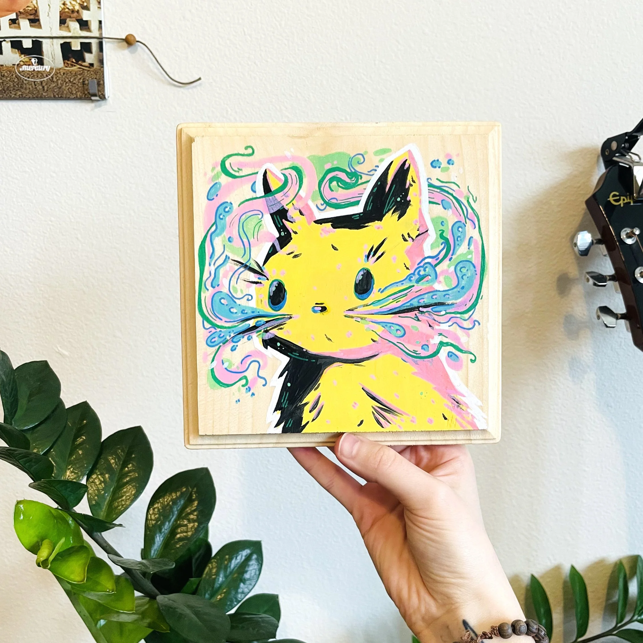 A wooden board with colorful artwork of a yellow cartoon character with black ears, surrounded by pink, green, and blue swirls, held by a person's hand against a white wall with a plant and jewelry on the wall in the background.