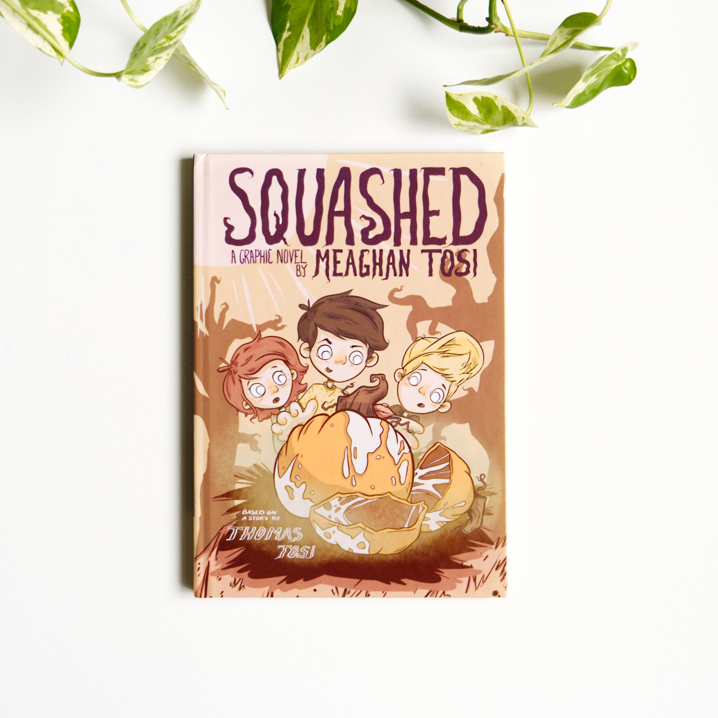 Close-up of a book titled 'Squashed' by Meaghan Tosi, with a colorful illustration of children on the cover, and green leafy plant on white background.