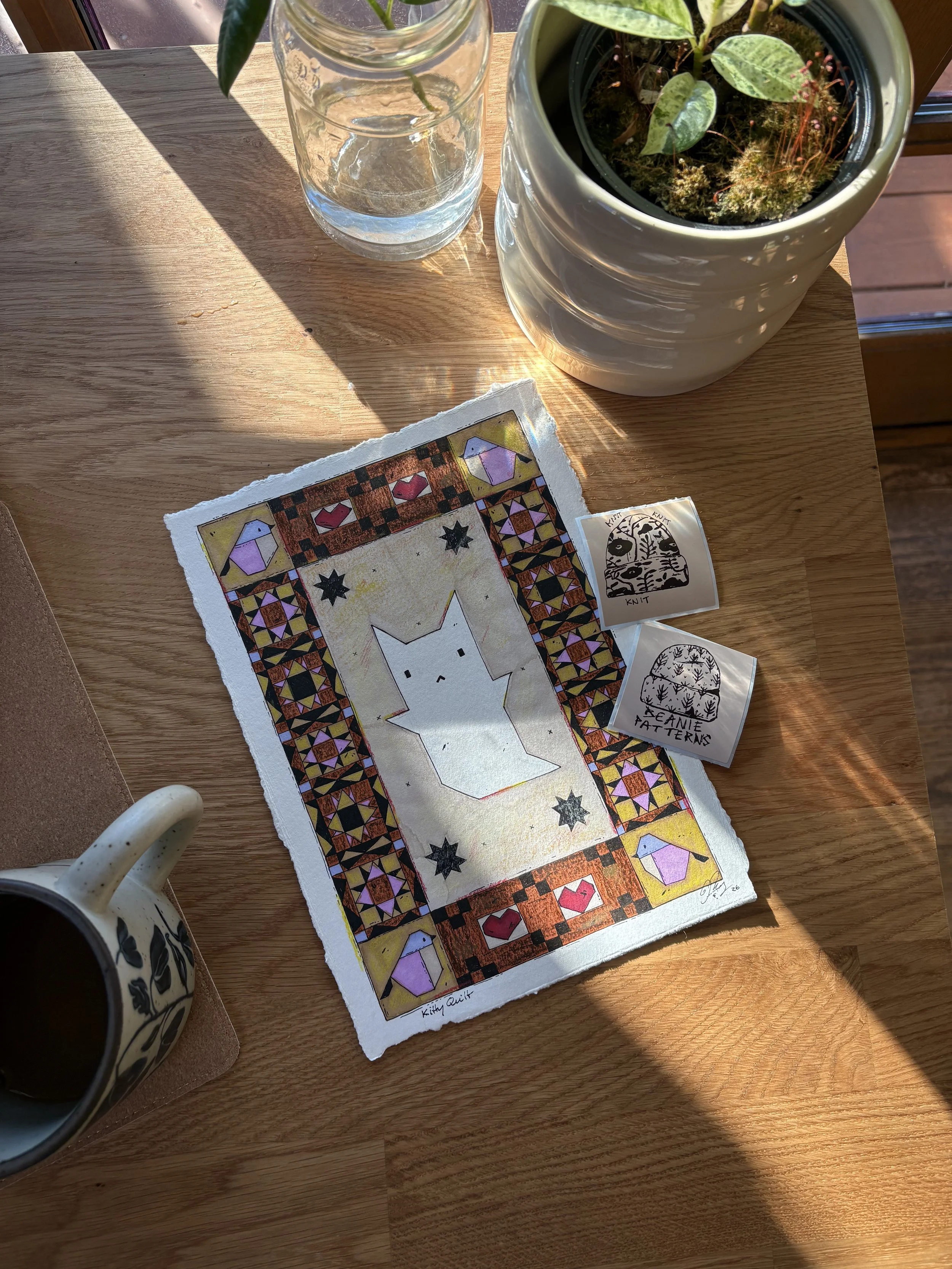 A wooden table with a potted plant, a glass of water, a black and white patterned mug, and a colorful quilt pattern print. The sunlight casts shadows on the table.