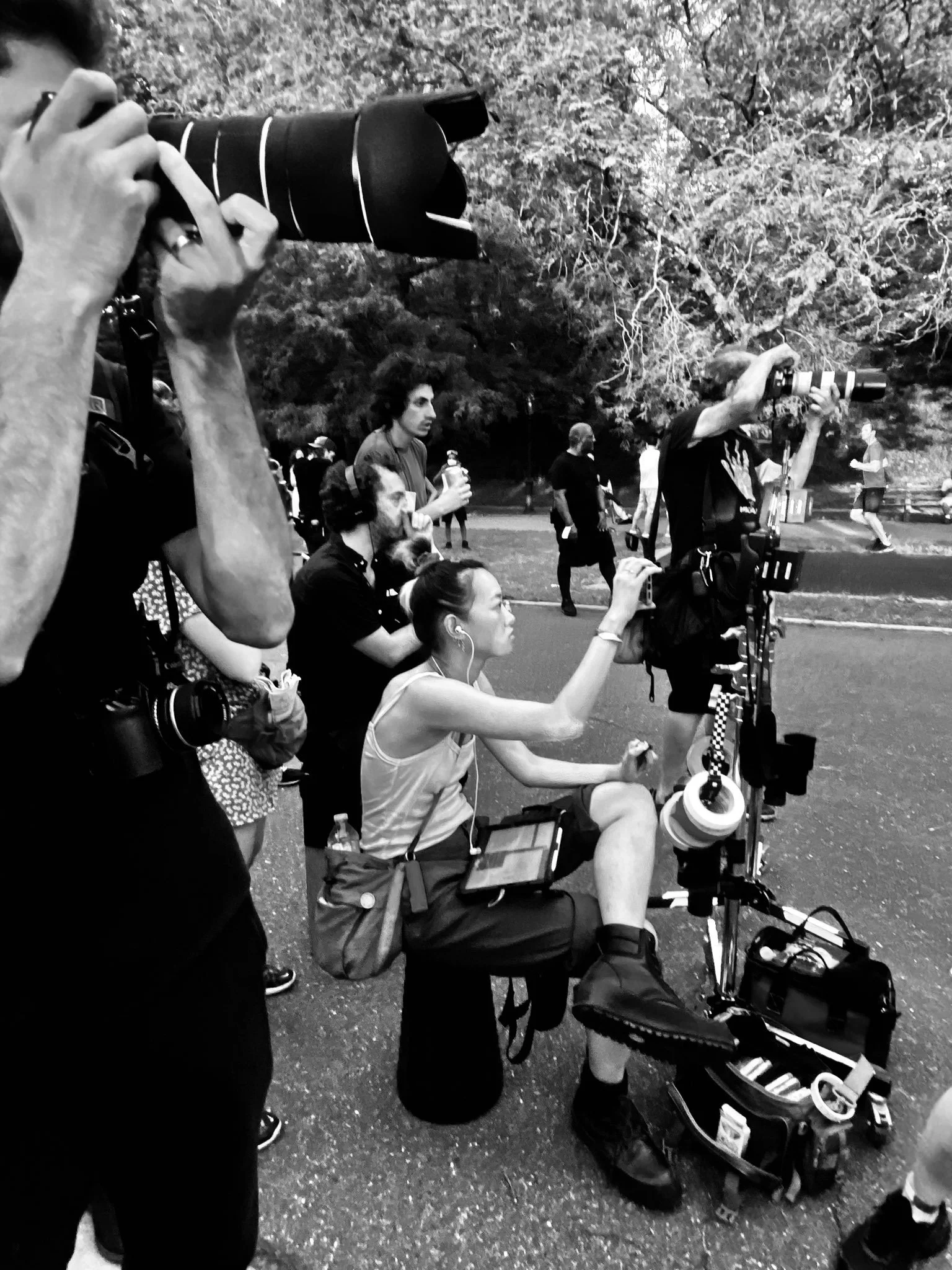 Group of people, some sitting and some standing, with two individuals using large professional cameras with telephoto lenses in a park with tall trees in the background.