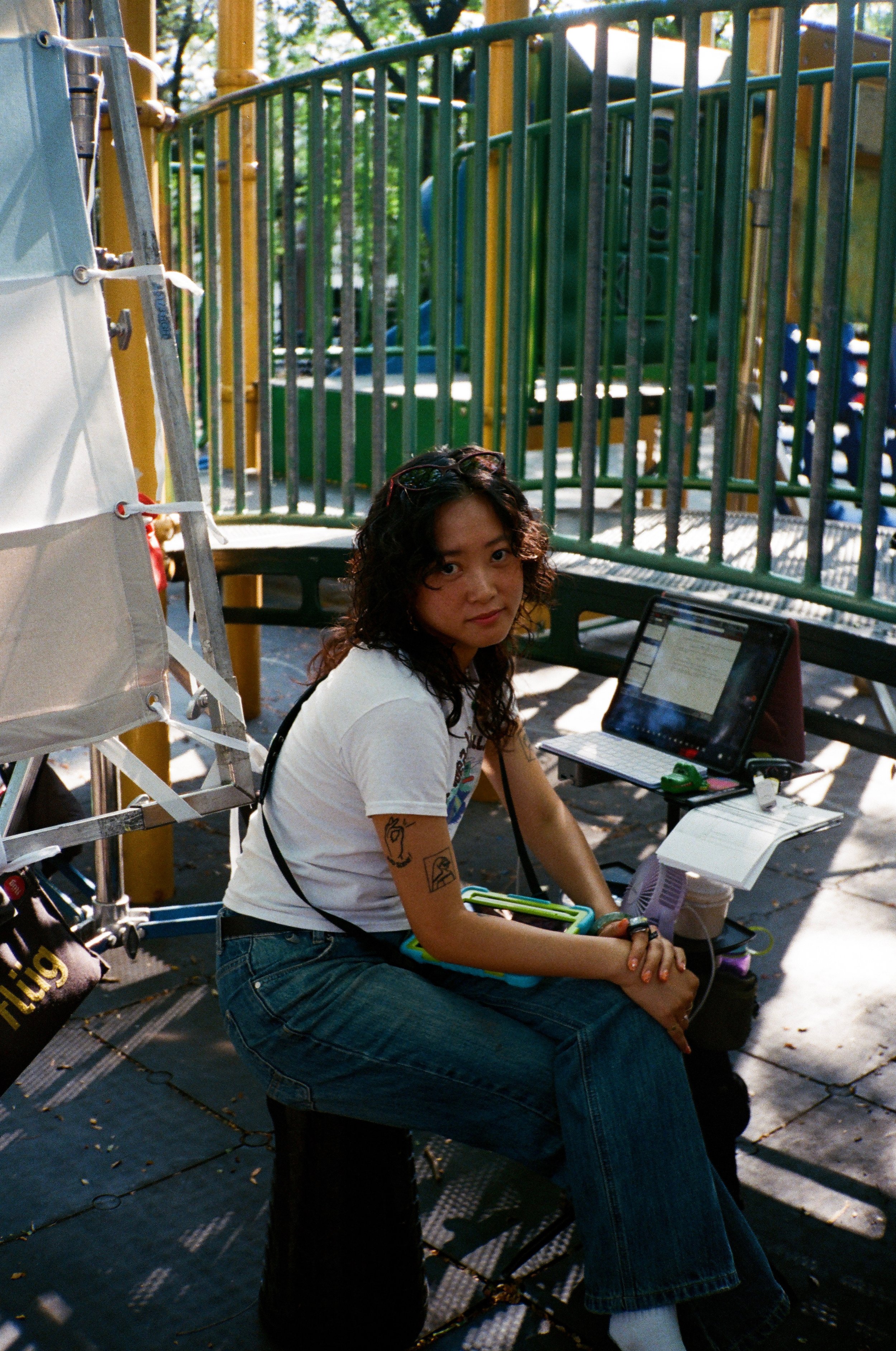 A young woman with curly dark hair sitting on a bench at an outdoor park, looking at the camera, with a laptop and various electronic devices around her. She wears a white t-shirt, blue jeans, and has tattoos on her arms. There is colorful playground equipment and a green fence in the background.