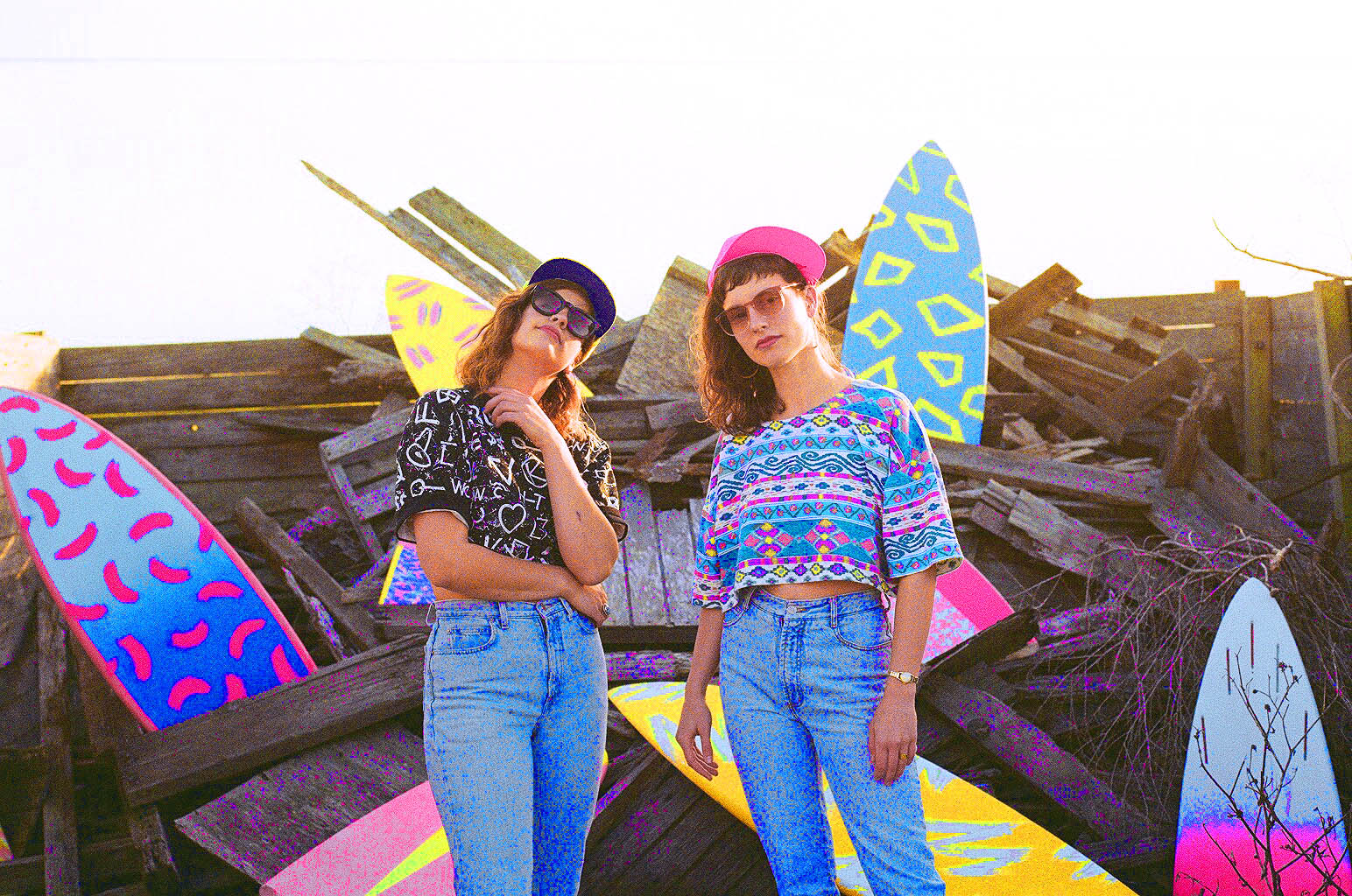 © EM (Emily Meehan) RadioActiveDaywear Two women in colorful, patterned clothing and large sunglasses standing in front of a pile of wood and painted surfboards with bright, geometric designs on a sunny day.