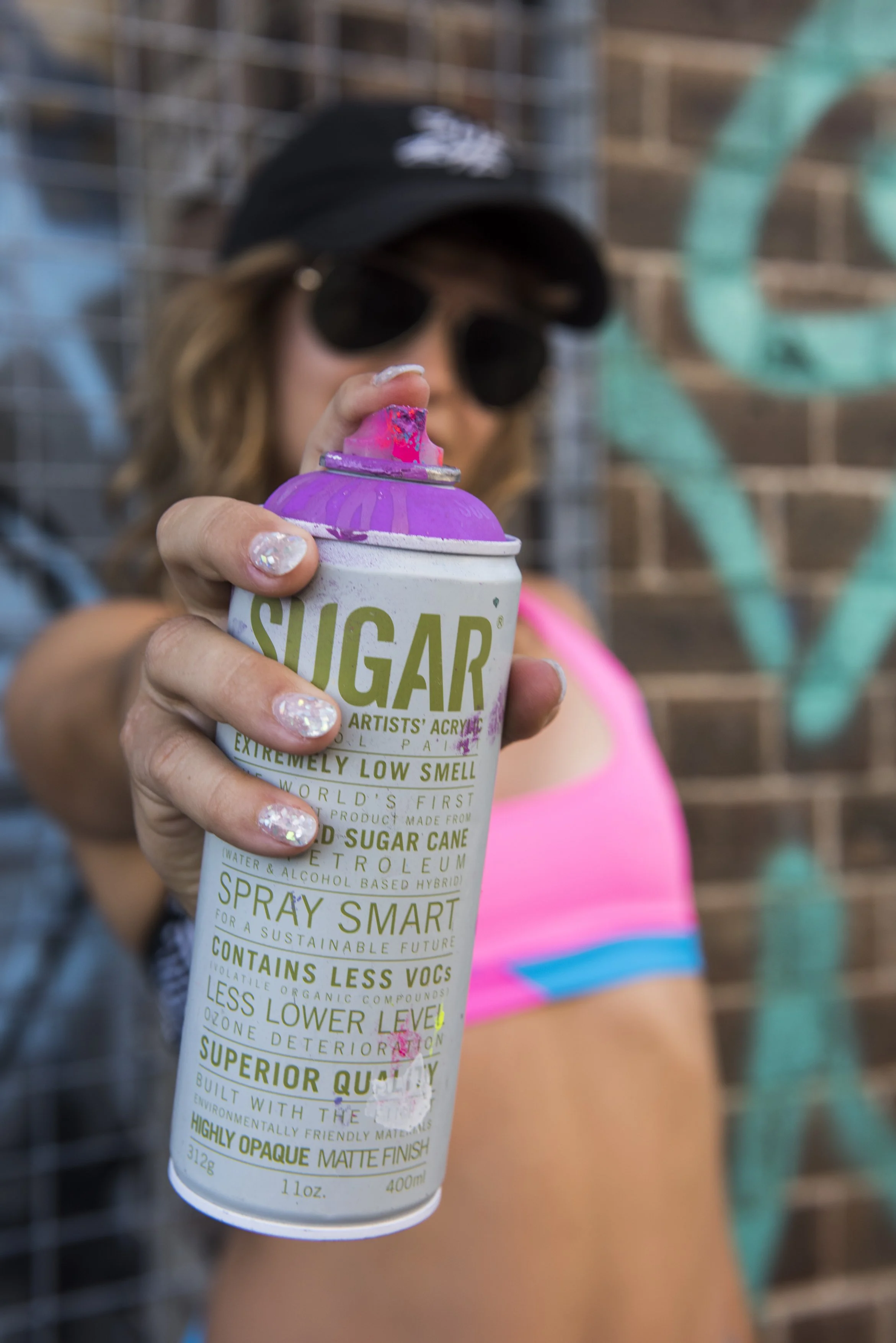 Person wearing black cap and sunglasses holding a can of spray paint with pink paint on the nozzle, standing in front of a brick wall with graffiti.