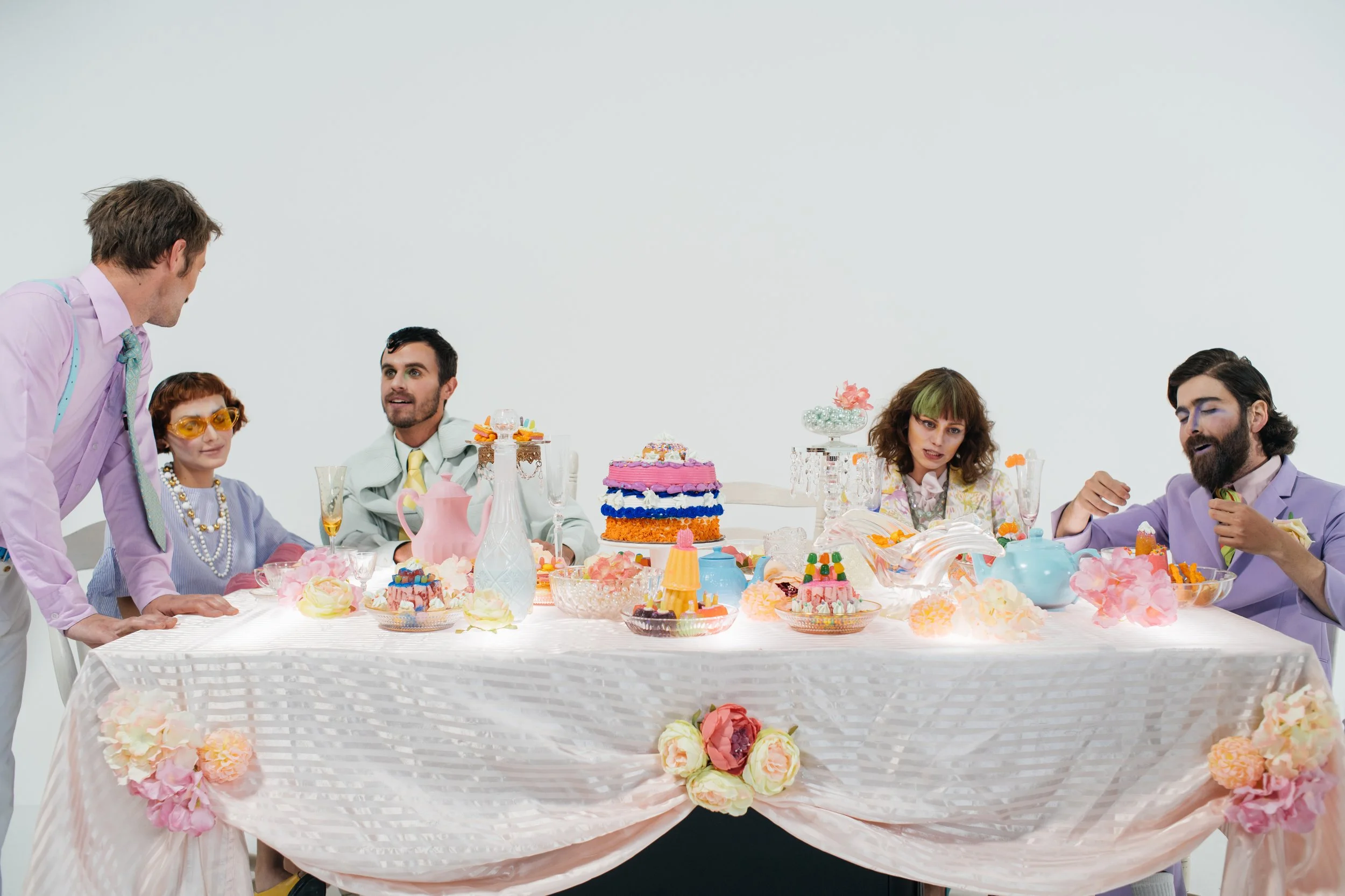 Five people at a decorated table with colorful cakes, candies, and flowers, engaging in conversation at a celebration.