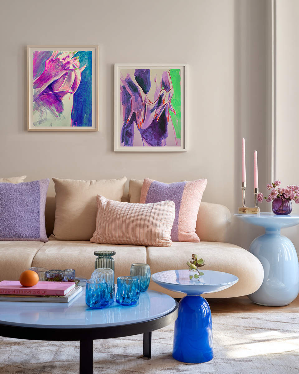 Living room with a beige sofa decorated with pastel-colored pillows, abstract purple and pink artwork on the wall, a coffee table with blue glassware, and a side table with pink candles and a purple vase with pink flowers.