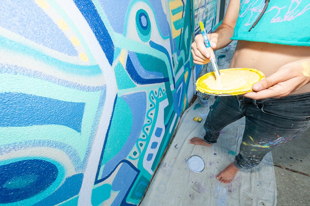 Person painting a colorful mural on a wall outside, holding a palette and brush.