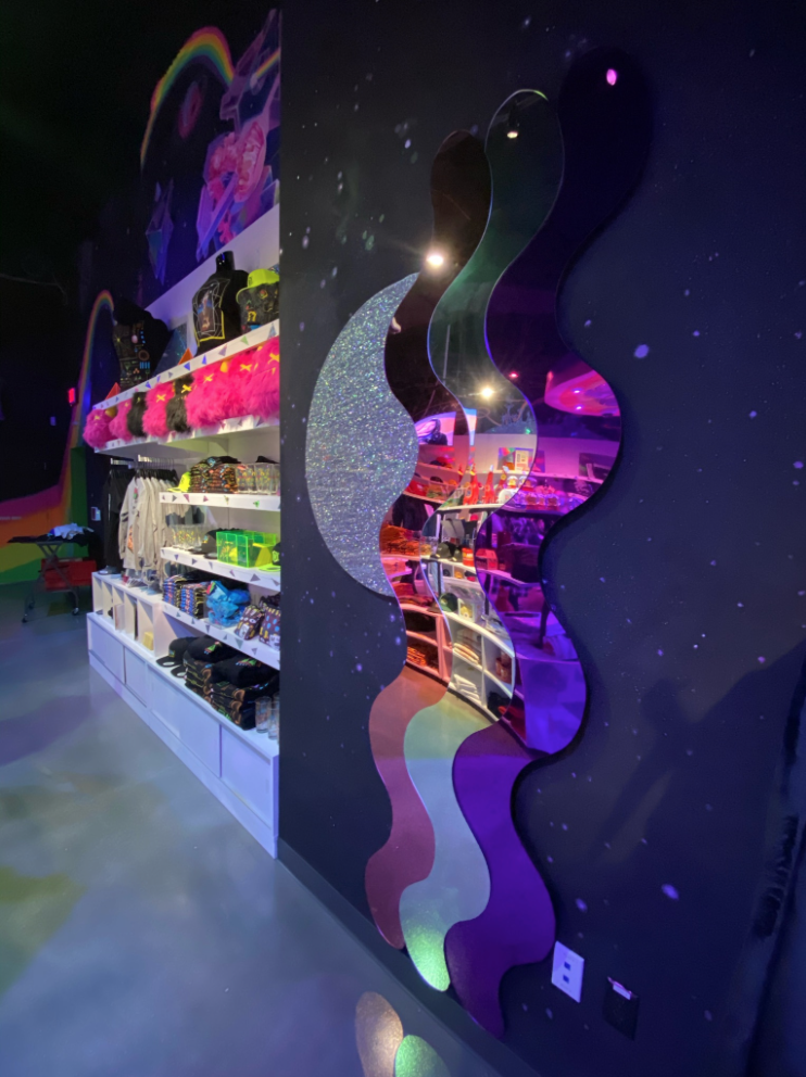 A store interior with colorful, wavy-shaped mirrors on a dark wall reflecting neon and galaxy-themed decor. Shelves display plush toys, bags, and clothing.