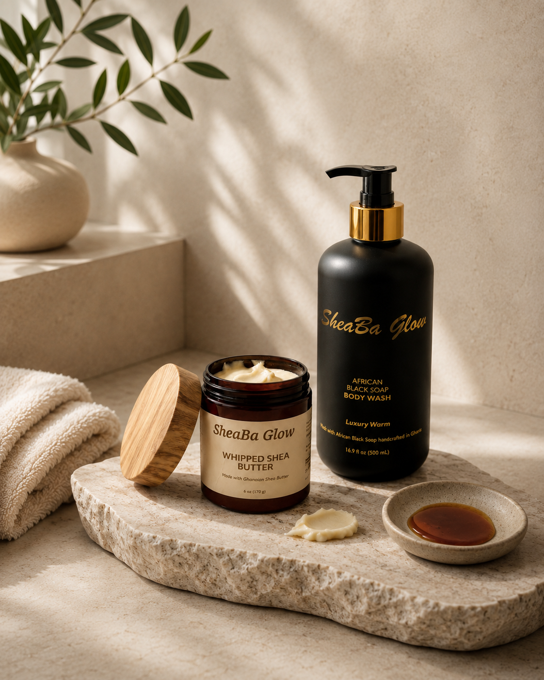 A flat lay of SheaBa Glow skincare products, including a black body wash bottle with gold accents and a jar of whipped shea butter, placed on a marble tray with a small dish of liquid, with a beige towel and green plant in the background.