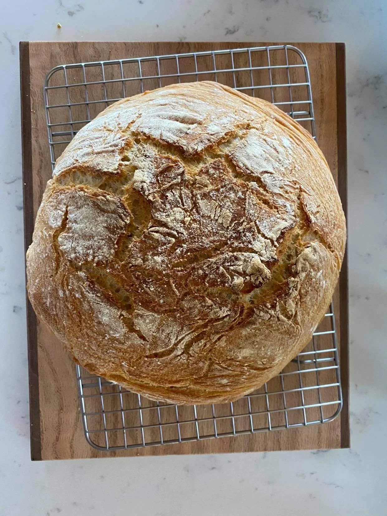 A round, freshly baked rustic bread loaf with a crusty surface, dusted with flour, resting on a cooling rack over a wooden cutting board.