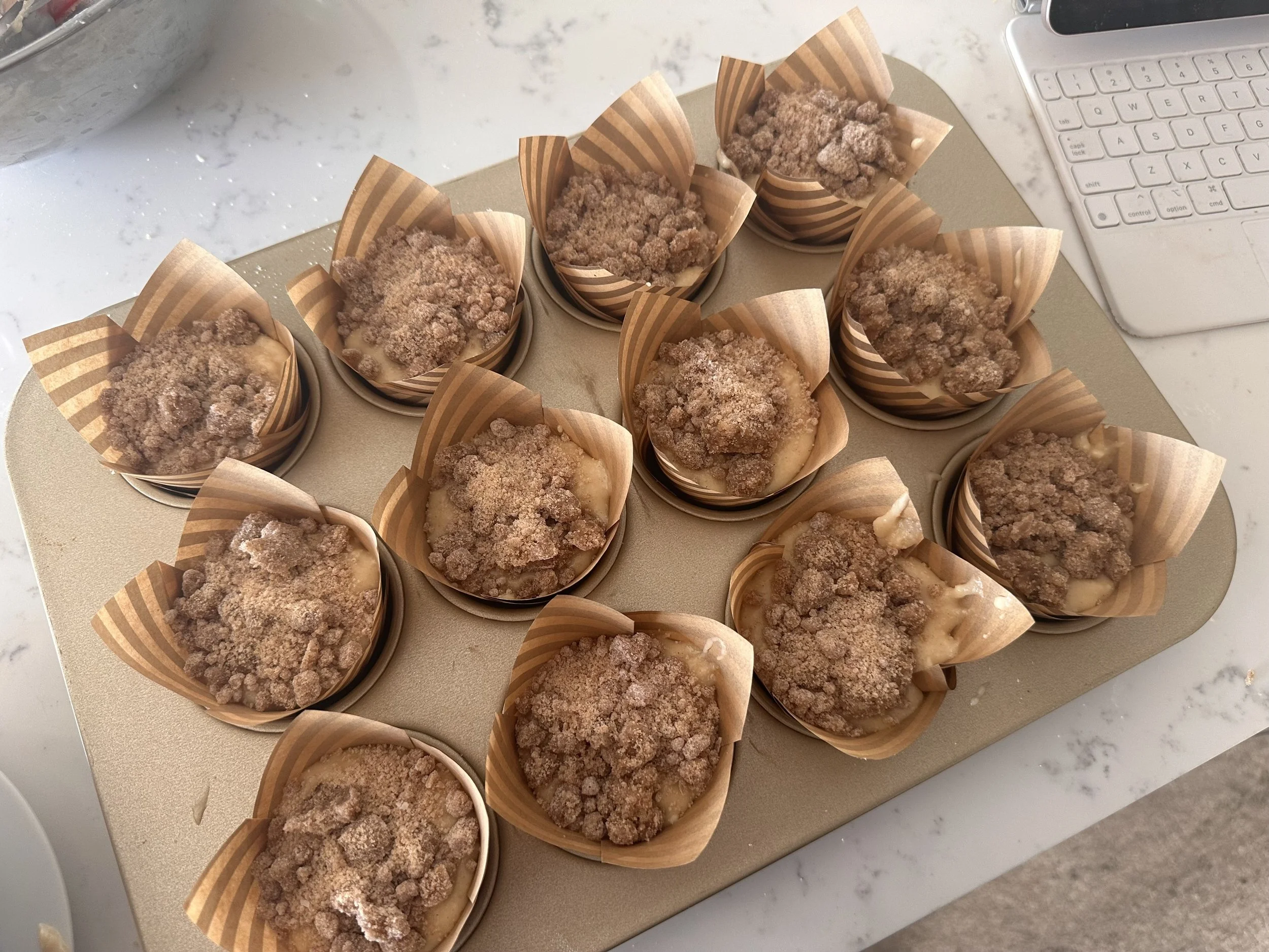 Multiple muffins topped with crumbled streusel in paper liners, arranged on a tray on a marble countertop near a keyboard and a metal bowl.