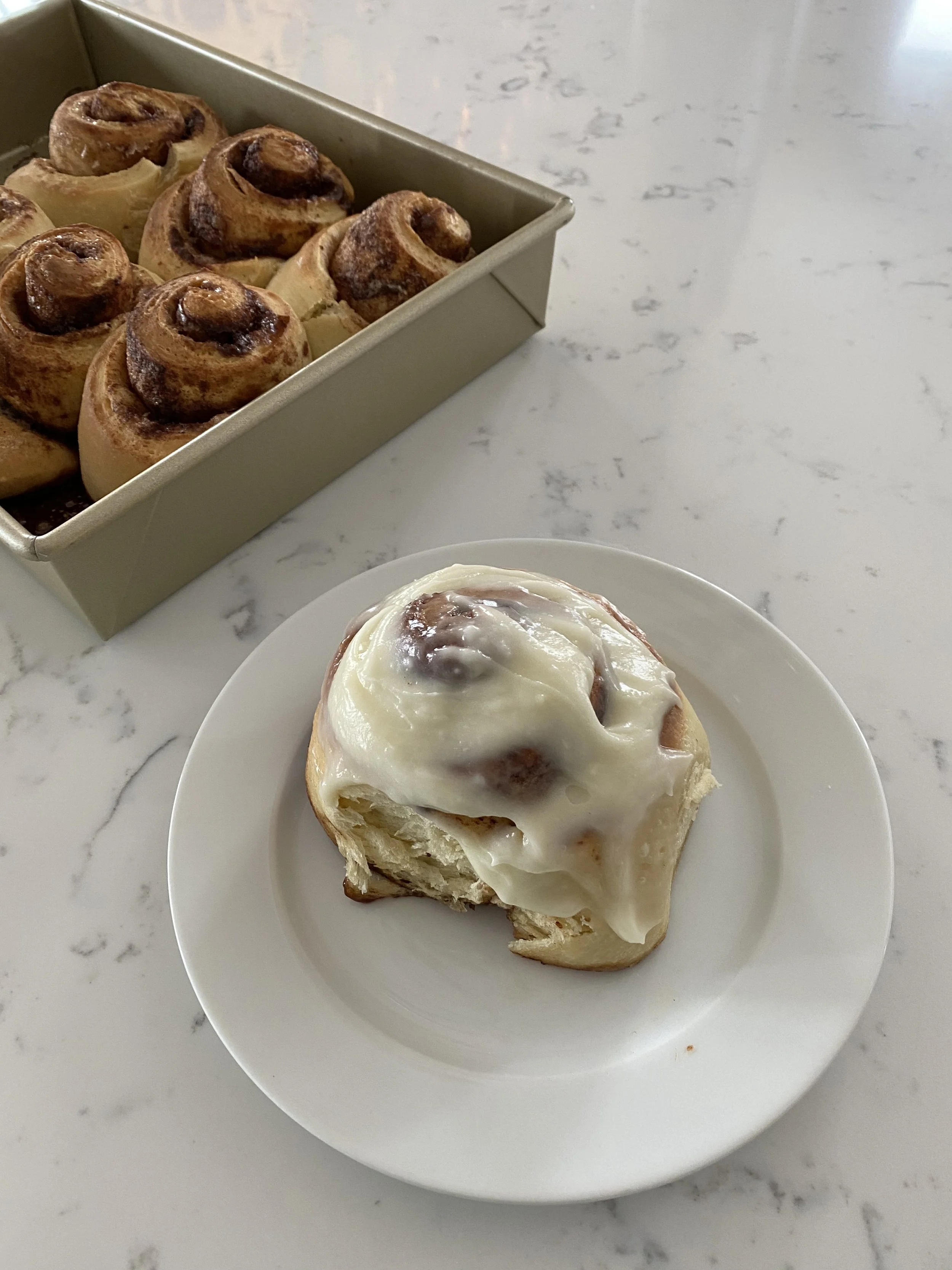 A cinnamon roll with melted white icing on top on a white plate and a tray of similar cinnamon rolls in the background.