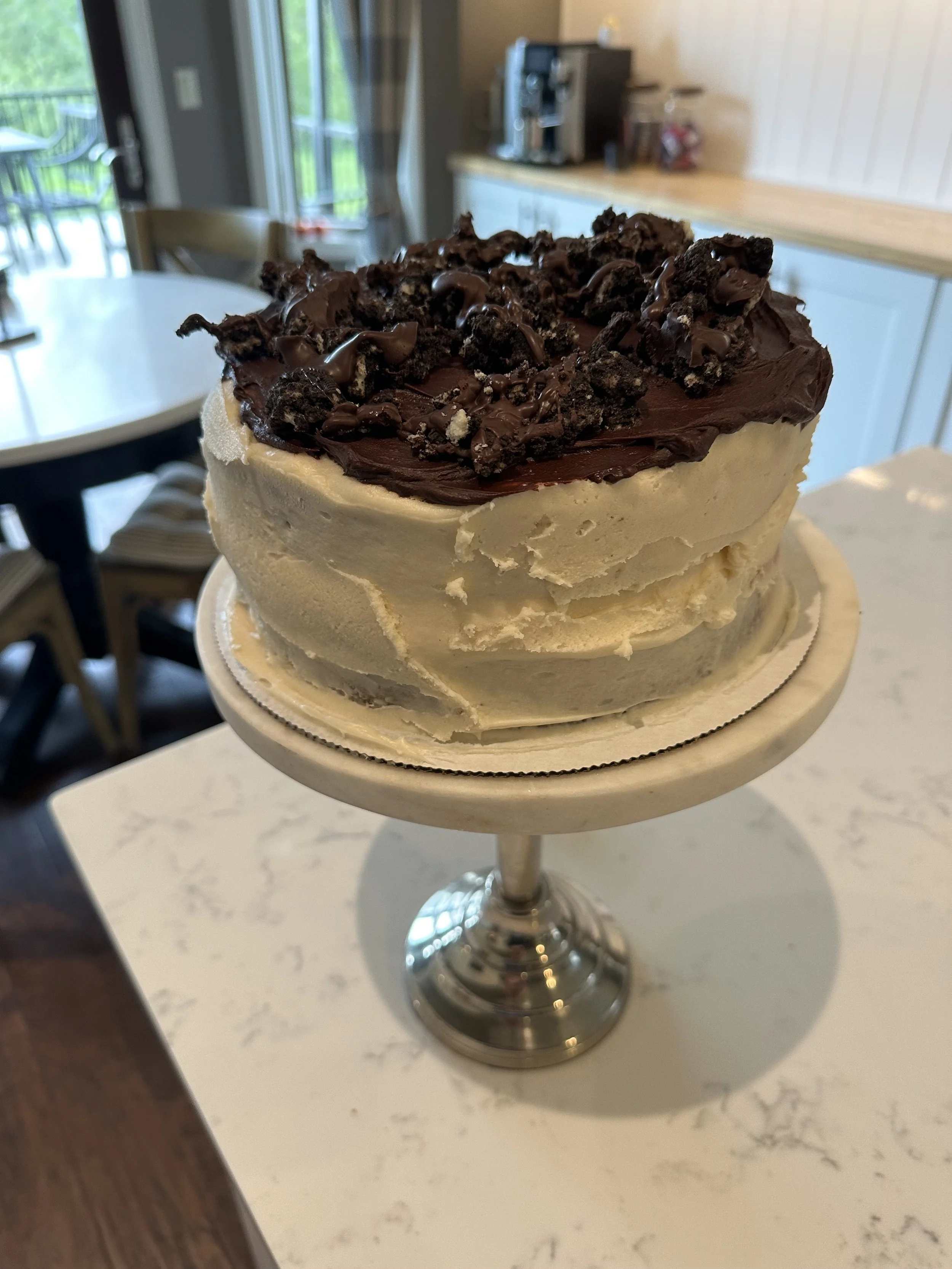 A cake with white frosting, topped with chocolate frosting and crumbled chocolate cookies, presented on a silver cake stand.