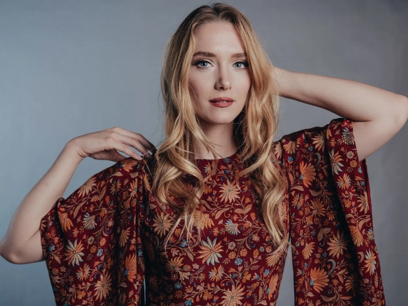 A young woman with long blonde hair, blue eyes, and fair skin, wearing a floral patterned dress with bell sleeves, posed with one hand behind her head and the other touching her shoulder, against a plain gray background.