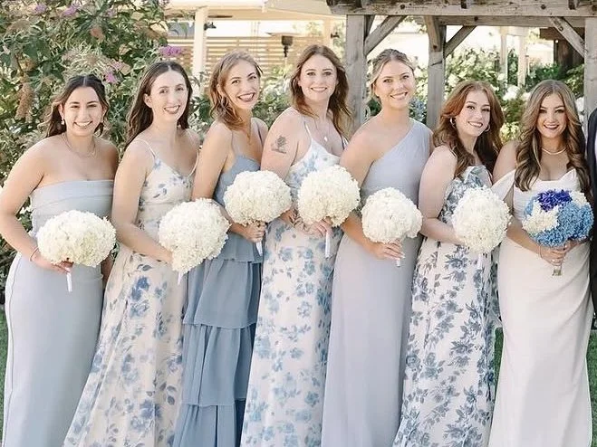 Seven women in pastel dresses holding white and blue flower bouquets, standing outdoors in front of a garden and wooden structure, smiling at the camera.