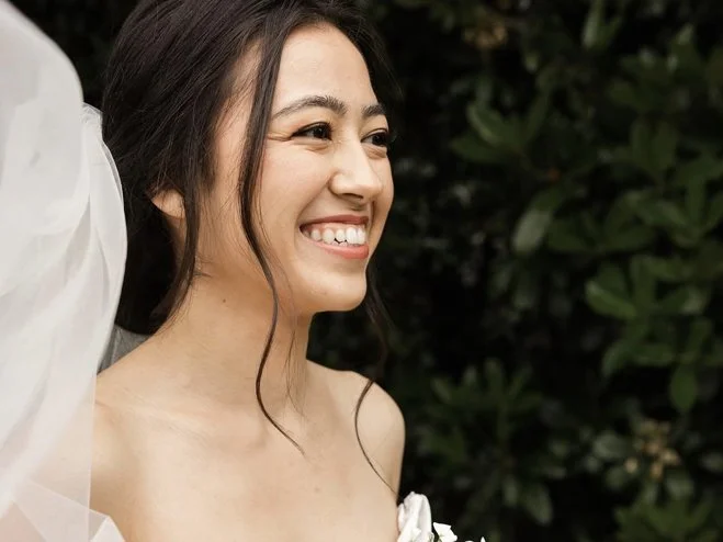 Smiling woman with long dark hair and a white dress, outdoors with green foliage in the background.