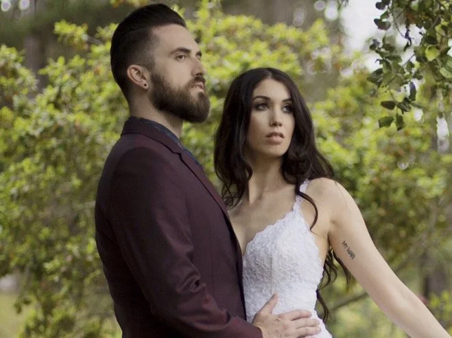 A couple standing outdoors in a natural setting, with the man in a dark suit and the woman in a white dress, both looking serious and contemplative.