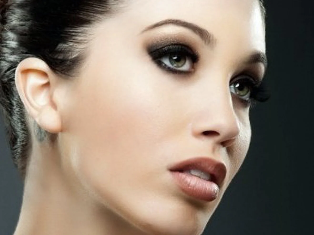 Close-up of a woman's face with makeup, dark eye shadow, and styled dark hair, against a dark background.
