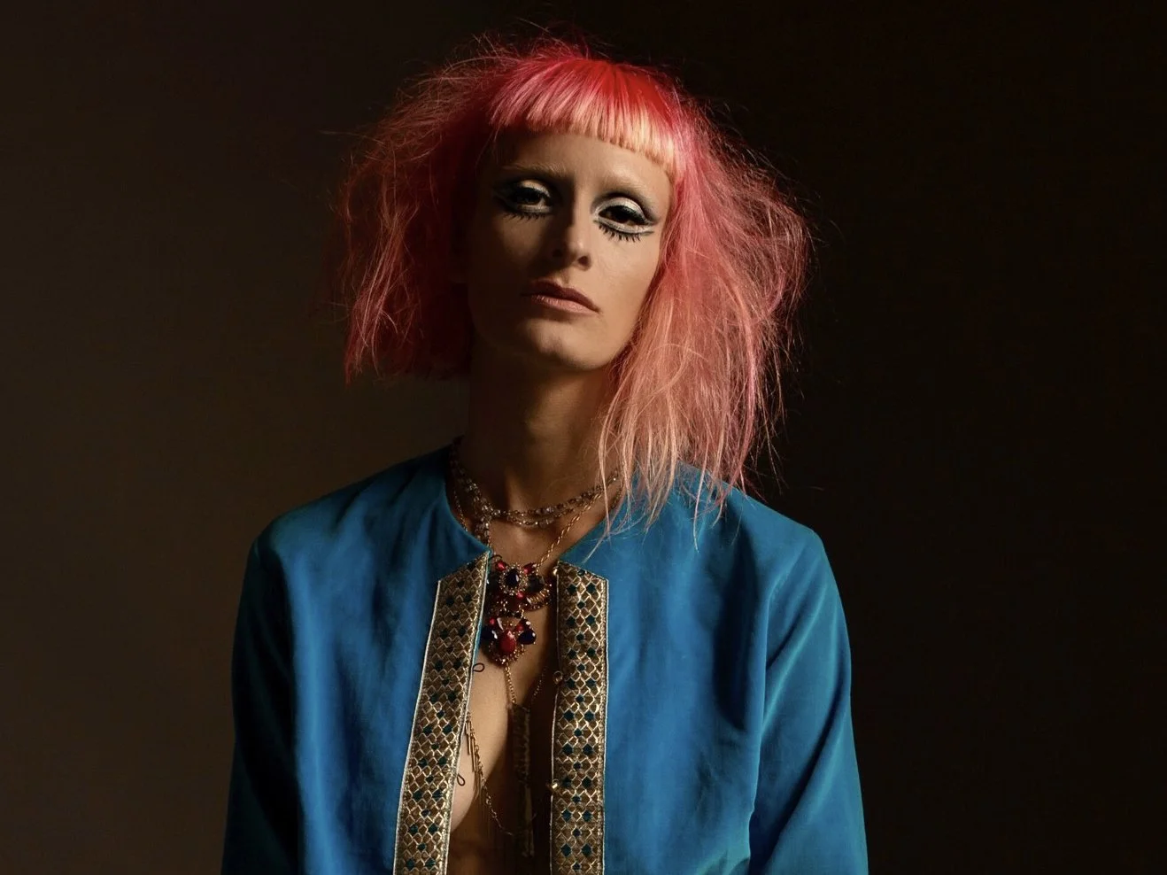 A woman with pink, messy hair, wearing a blue jacket with gold embroidery and layered jewelry, posing against a dark background.