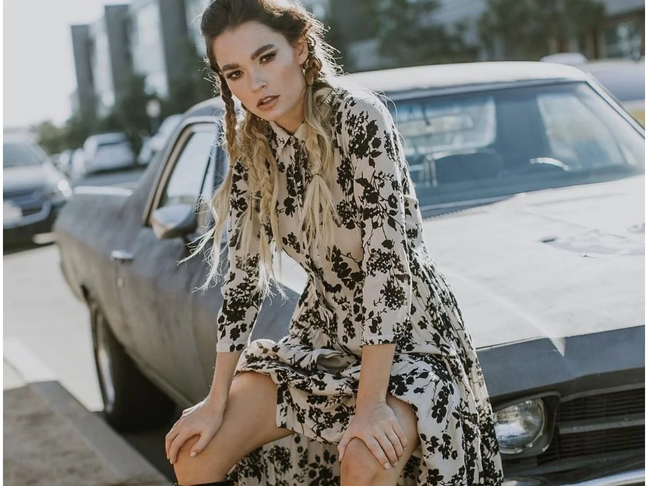A woman with braided blond hair in a floral dress sitting on the hood of a vintage gray car in a parking lot.