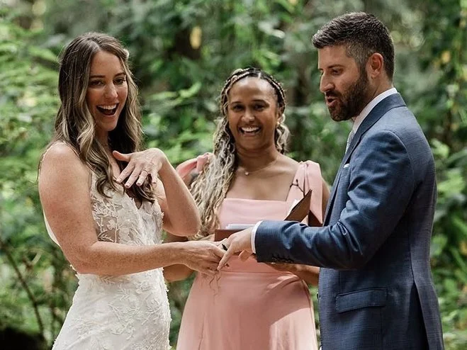 A wedding ceremony takes place outdoors with two women and a man. One woman is wearing a white wedding dress and the other in a pink dress, while the man is dressed in a blue suit. They are exchanging rings and smiling.