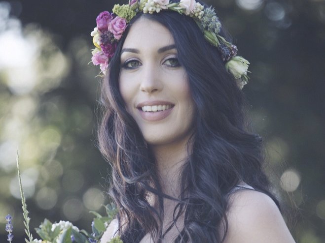 A young woman outdoors with long dark hair, wearing a floral crown made of pink, purple, and white flowers, smiling at the camera with a blurred nature background.