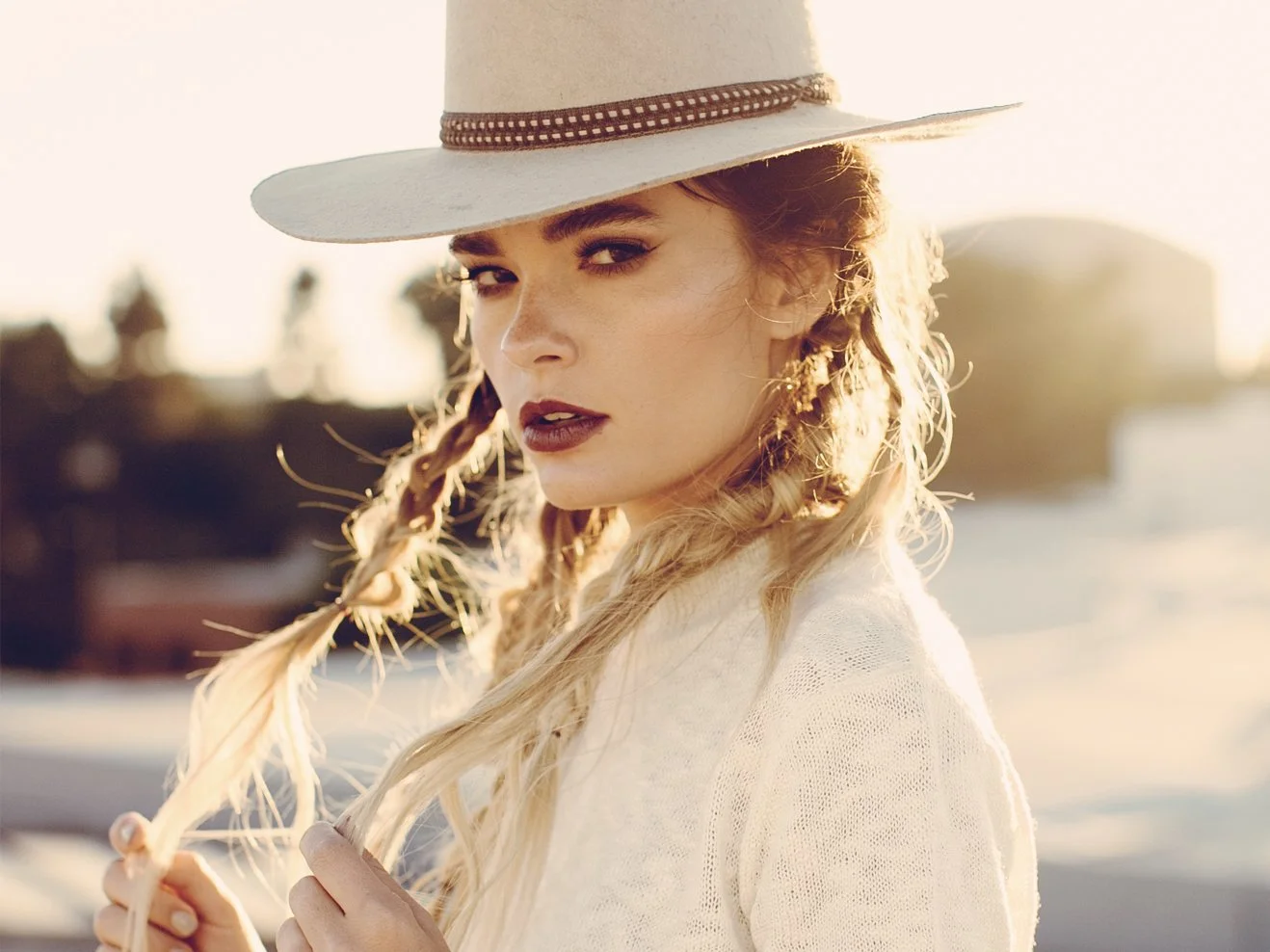 Close-up of a woman with blonde hair, wearing a wide-brimmed hat and a white sweater, standing outdoors during sunset.