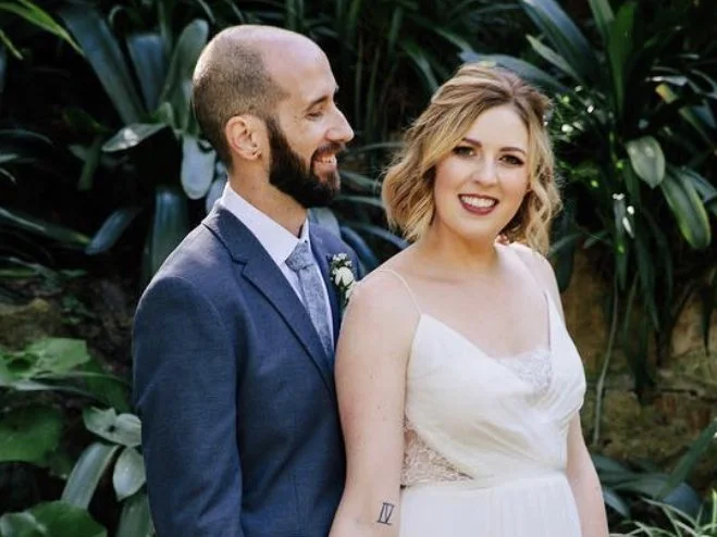 A happily married couple at their wedding, standing outdoors among green plants. The groom has a beard and is wearing a blue suit, while the bride has blonde wavy hair and is dressed in a white wedding gown, smiling at the camera.