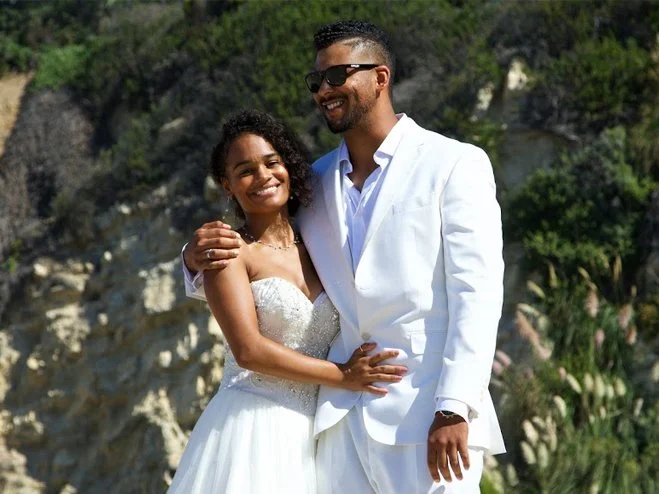 A happy couple in wedding attire outdoors with greenery and rocks in the background.