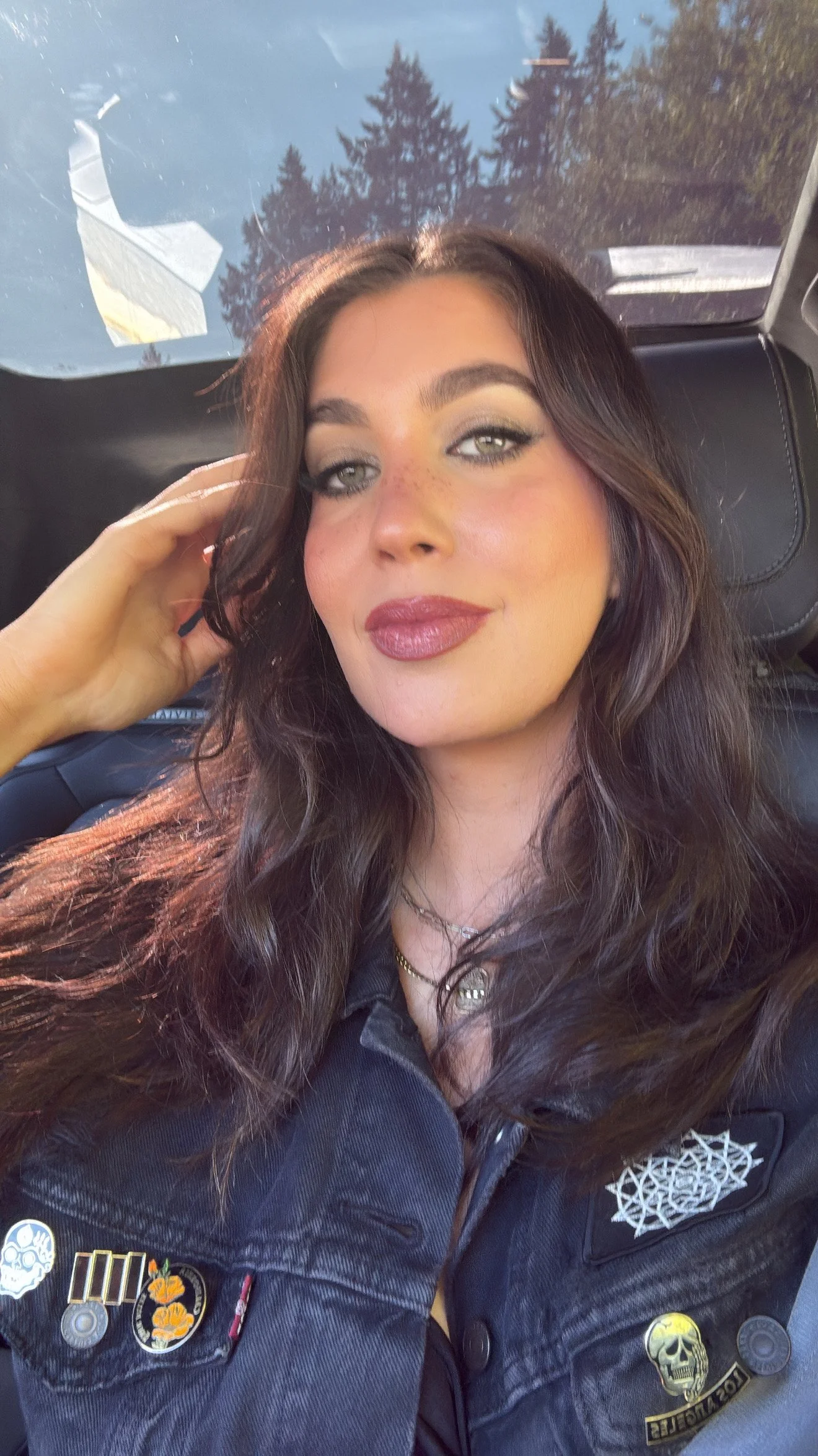 A woman with long wavy brown hair and makeup, wearing a black shirt and a black jacket adorned with various pins and patches, sitting inside a vehicle with trees and a blue sky visible through the window behind her.
