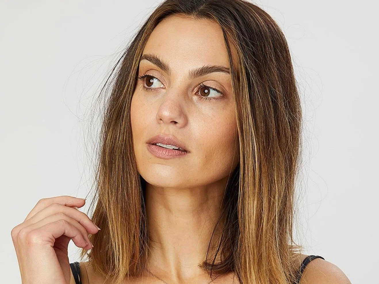 Close-up portrait of a woman with straight, shoulder-length brown hair, light makeup, and a neutral expression, looking slightly to her left against a plain gray background.