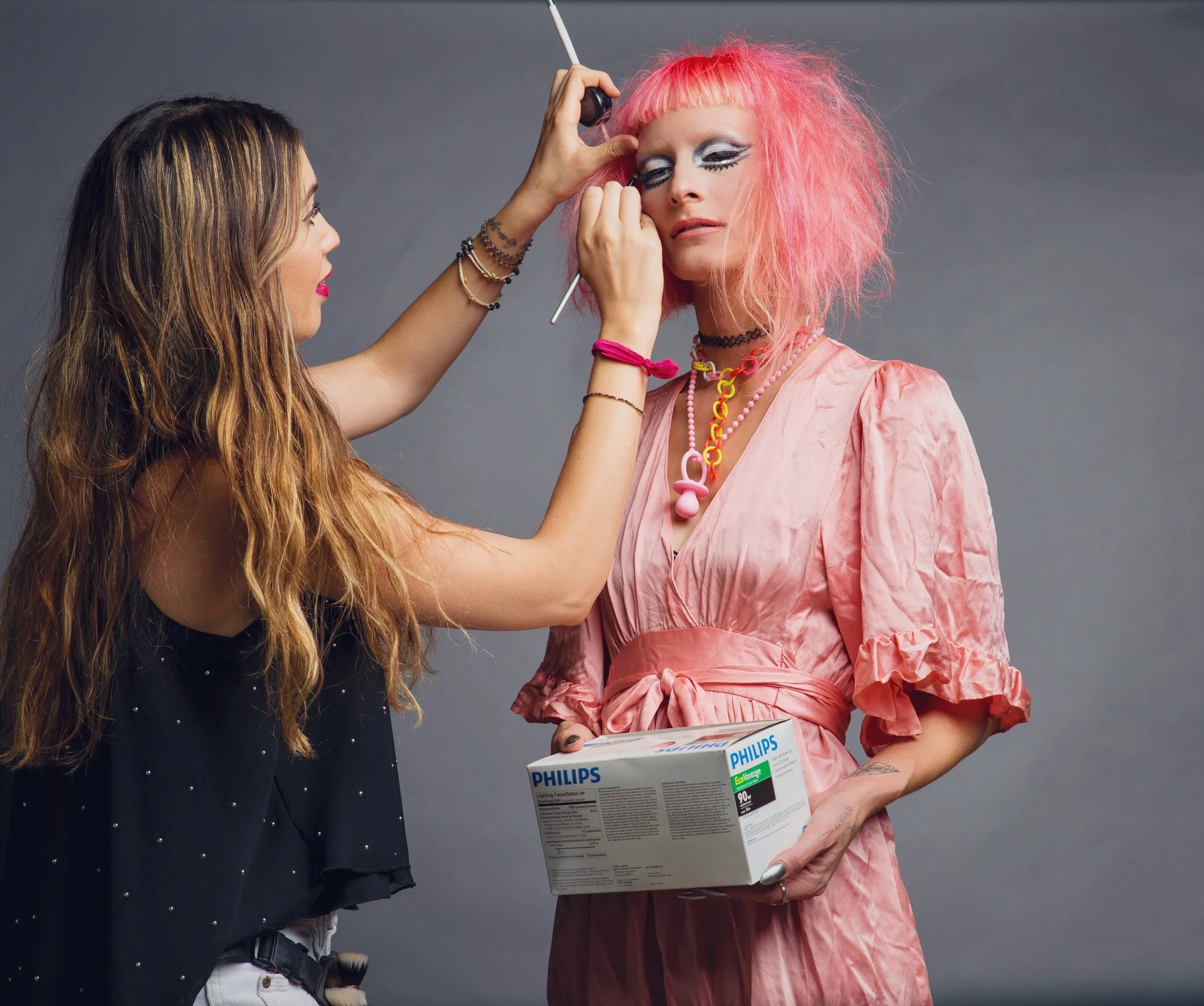 An artist is applying makeup to a woman's face during a photo shoot, with the woman holding a box of Philips light bulbs.