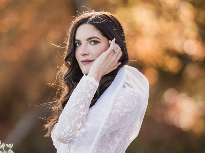 A young woman with long dark hair wearing a white lace dress standing outdoors during autumn, with warm blurred fall foliage in the background.