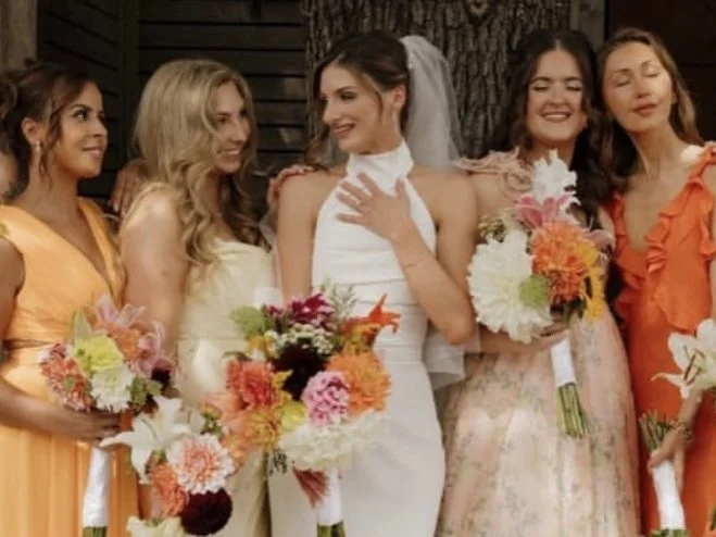 Group of five women at a wedding, holding colorful flower bouquets, standing outdoors in front of a tree and rustic wooden wall.