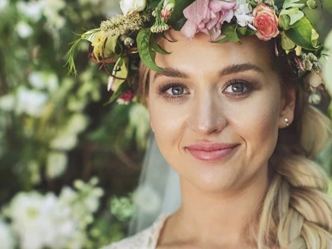 A woman wearing a flower crown made of pink and white flowers, smiling with a natural outdoor background.