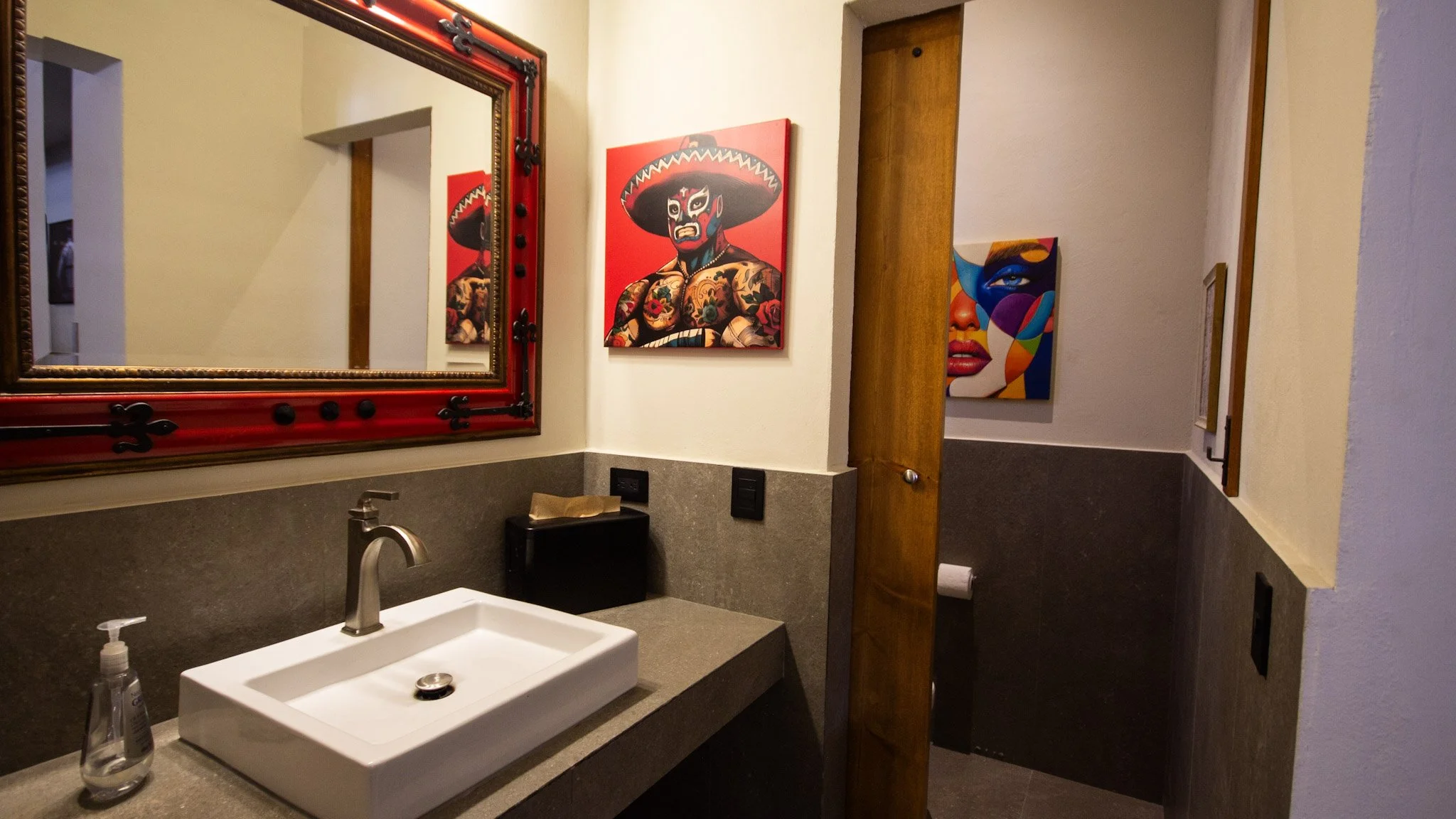 A bathroom with a white sink, a soap dispenser, and a mirror with a decorative red and black frame. Artwork featuring vibrant, tattoo-style illustrations of a woman with a hat and tattoos on the wall. A wooden door to the right and a partial view of 