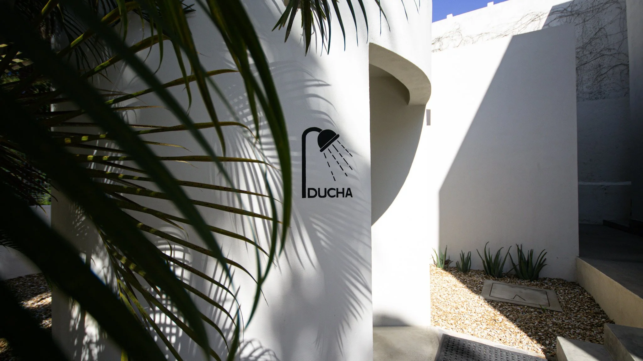 Modern outdoor shower area with white walls, a sign with a shower head icon and the word 'DUCHA', plants, gravel ground, and shadows cast by nearby structures.