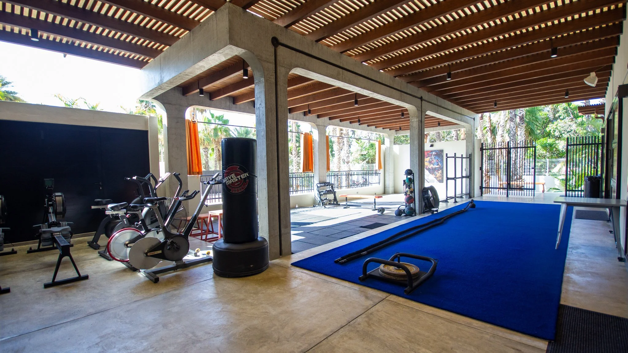 Indoor gym area with exercise equipment, punching bag, and weights, opening to outdoor space with palm trees and a fence, under a wooden slat ceiling.