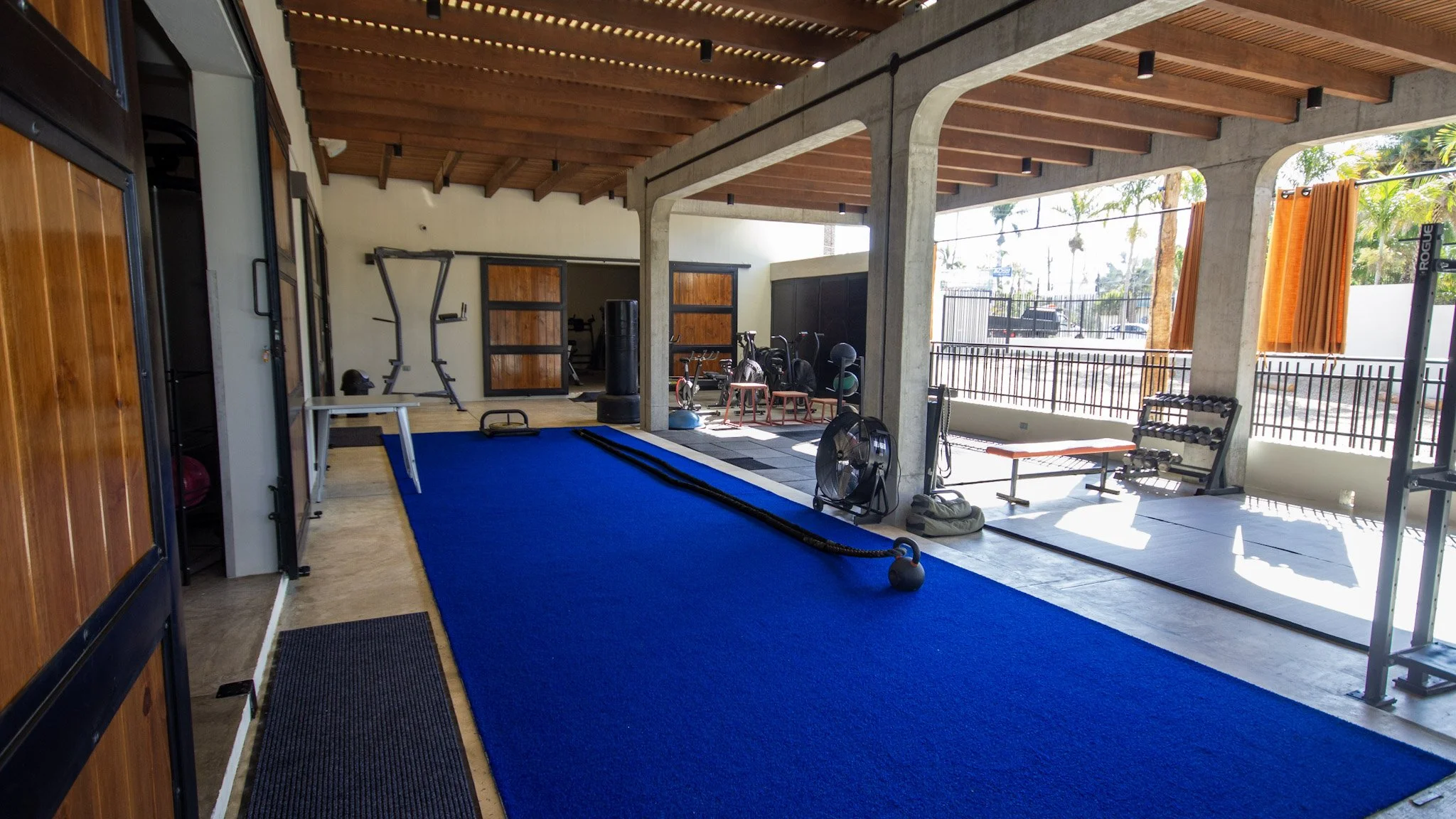 Indoor gym space with workout equipment, including free weights, a punching bag, and resistance bands, with large open windows and orange curtains outside.