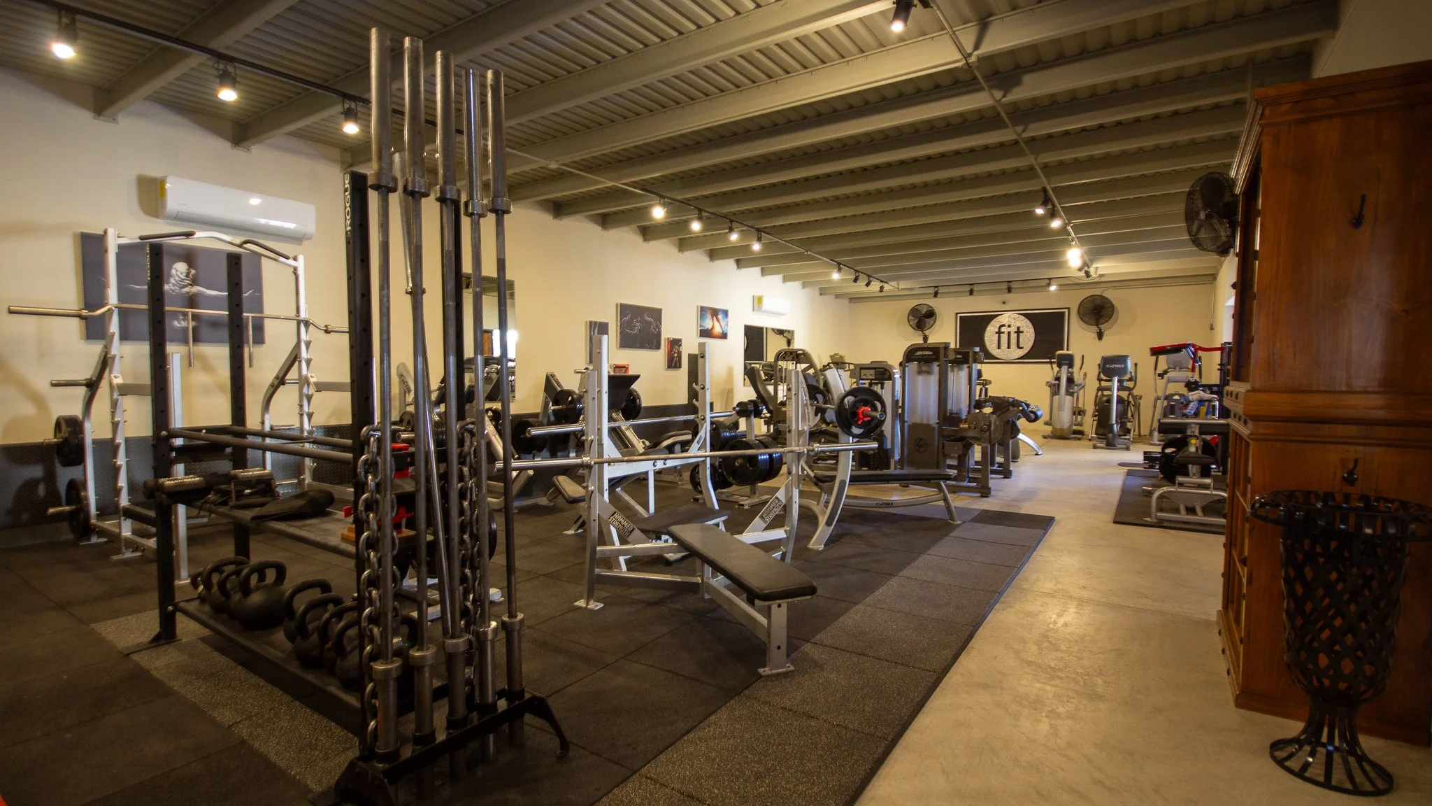 Interior of a gym with various workout equipment, including weightlifting racks, benches, and cardio machines. The space has high ceilings with track lighting and wall fans, and a sign that reads 'fit' in the background.
